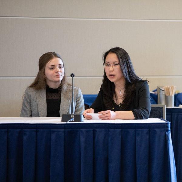 two first-year students sit at a table with microphone between them. Table has blue draping