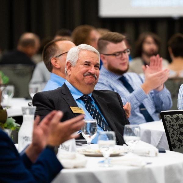 Paul Plotkowski is seated at a table smiling as others clap for his accomplishments