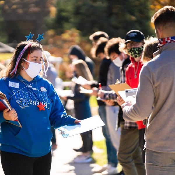 A student in a blue GVSU sweatshirt and holding clipboards hands a clipboard to a student waiting in a line to vote.