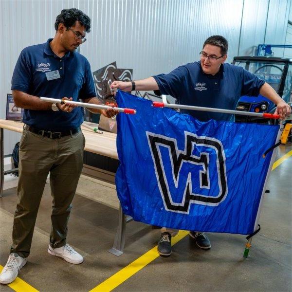 two men stand behind a GV flag while placing a pole through the flag