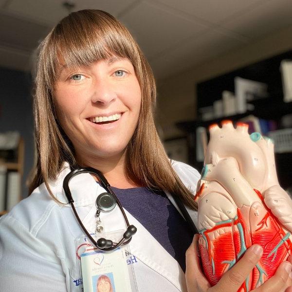 Cari Bellicini is in a white medical coat holding a plastic heart model