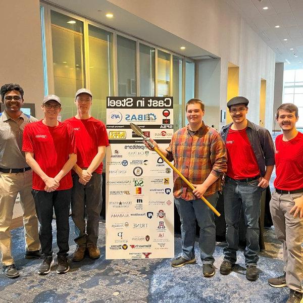 six people standing, one holding spear, next to vertical poster of Cast in Steel teams with university logos.