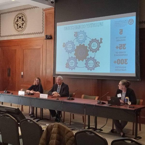 three people seated at a table, projection screen in back shows GVSU slide of gears, discussing PCEC's impact in the community